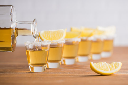 Row of glass sots with golden tequila and slices of lemon on wooden table with white wall on blurred background