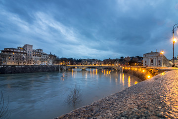 Fototapeta premium Long exposure view of the Tiber river with bridge in the background on a rainy autumn sunset with light set and reflection, Rome, Italy. Top view.