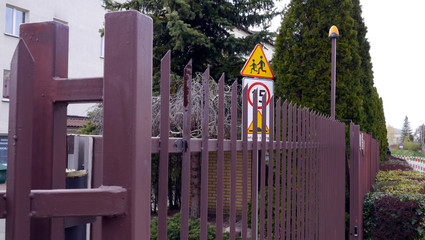 Brama wjazdowa na osiedle. Znaki ostrzegawcze "uwaga dzieci" oraz ograniczenie prędkości do 15. Osiedle mieszkaniowe wśród drzew, obok uliczka osiedlowa. Wjazd, wyjazd