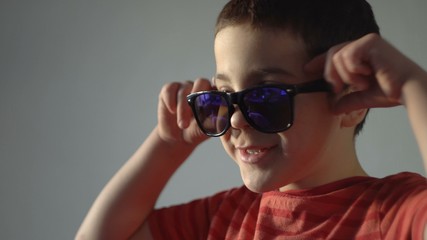 A boy stands against a gray wall and laughs. There are dark glasses on the face. Dark hair. The sun at sunset. Age 6 years. Close-up.