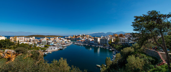 Agios Nikolaos City and Voulismeni Lake. Panorama of the beautiful of Agios Nikolaos. Panoramic view sea, lake with trees, sky and reflection on water. Agios Nikolaos, Crete, Greece.