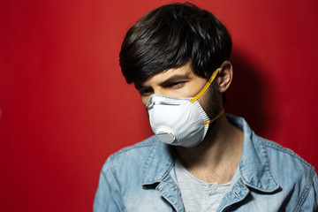 Studio portrait of young sick guy in denim jacket wearing white medical flu mask against coronavirus, isolated on red background.