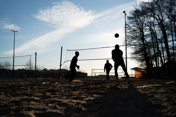 Silhouettes of people at sunset playing volleyball. Ball in the air, tense postures of people..A game of beach volleyball in cold weather in clothes.