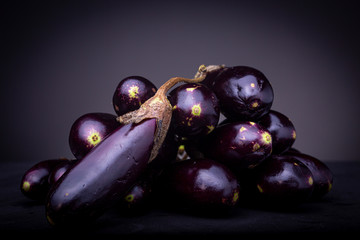 Pile of small colourful vibrant purple baby eggplant aubergine vegetable with one in front. Studio low key food image on black surface and dark grey background.