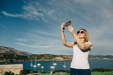 Happy blonde woman tourist taking selfie on top of a hill overlooking the sea and mountains