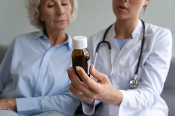 Close up of young woman doctor hold medicine bottle prescribe liquid syrup to sick senior grandmother patient, female nurse explain meds prescription to mature grandma, elderly healthcare concept