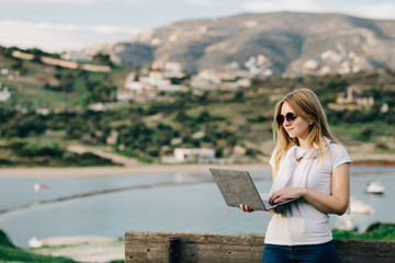 Young blond girl freelancer in glasses working on a laptop with sea and mountain views