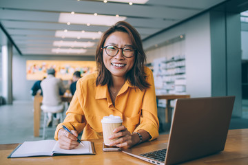 Portrait of happy Asian woman smiling at camera during studying process in university campus feeling excited from exam preparation, emotional female in trendy spectacles e learning in coffee shop