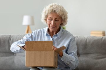 Excited mature woman sit on couch in living room open cardboard box shopping online at home, happy...