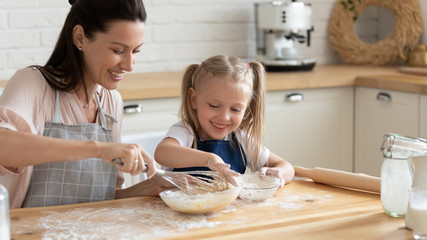 Happy little girl child helper cooking stir whisk eggs prepare dough for cookies with young mom, smiling mother teach small daughter make pancakes or pie, baking family recipe breakfast together