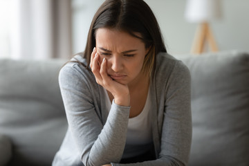Upset young woman sit on couch in living room lost in thoughts feel lonely and distressed, sad...