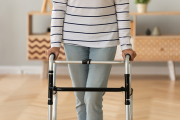 Close up of sick disabled senior woman standing making steps holding walker in rehabilitation center, mature ill handicapped old lady walk with walking frame in clinic, physiotherapy concept
