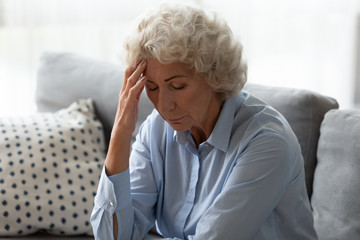 Fototapeta na wymiar Sick senior woman sit on sofa at home feel distressed suffer from migraine or headache, ill mature female rest on couch touch head have dizziness from high blood pressure, elderly healthcare concept