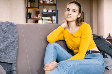 happy young girl with a cute face in an orange sweater and jeans is resting while sitting on a couch