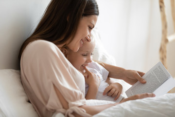 Close up of smiling mom and little daughter relax in cozy bed read funny interesting children book together, happy mother and small preschooler girl child enjoy fairytale rest in comfortable bedroom