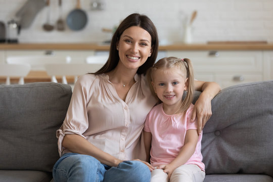 Portrait of happy young woman sit together hug cuddle on cozy couch with little daughter in living room, smiling loving mother and girl child embrace relax on comfortable sofa looking at camera