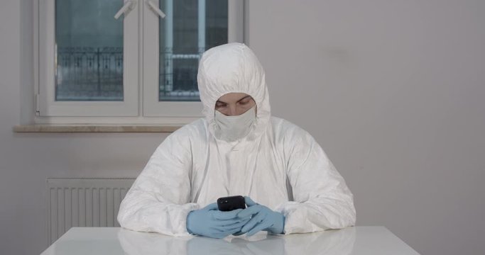 Portrait of young male person stitting behind white dining desk in kitchen in protective suit reading news on his phone during pandemic coronavirus quarantine time 4K