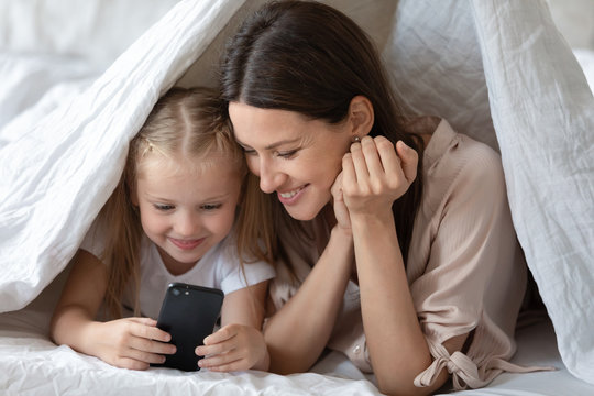 Smiling young mother and small preschooler girl child hide under blanket have fun using smartphone watching cartoons together, excited happy mom and little daughter play on cellphone in bedroom