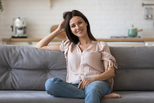 Portrait of smiling young woman sit relax on comfortable couch enjoy weekend in cozy home, happy millennial girl rest on sofa in living room looking at camera posing, renting, ownership concept