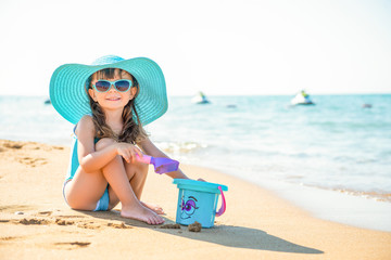 Little child girl fashionista in a blue wide-brimmed hat and sunglasses sits on the sand with a bucket on the seashore