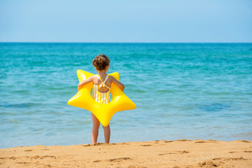 A little child girl 3 years old stands with a yellow swimming ring in the form of a starfish and looks at the sea. Back view