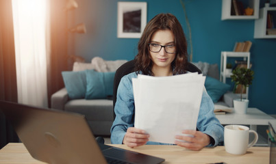 Focused young woman working at home looking at papers sitting at desk next to laptop computer