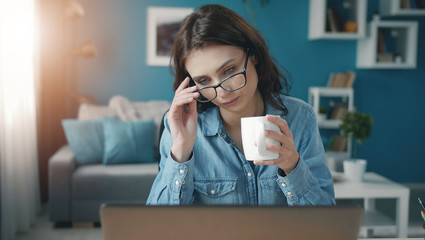 Focused young woman holding cup and touching glasses looking at laptop screen, livingroom background