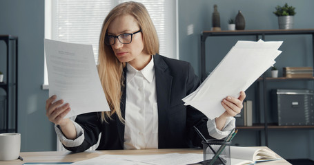 Focused blond business lady sitting at office table holding lots of docs reading paper reports