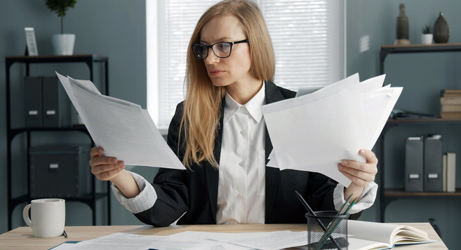 Concentrated blond business woman sitting at office desk holding heaps of documents, paper reports