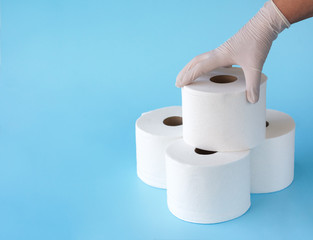  Female hand in medical gloves takes a roll of toilet paper from a folded tower of rolls on blue background. Toilet paper crisis due to coronavirus  quarantine.