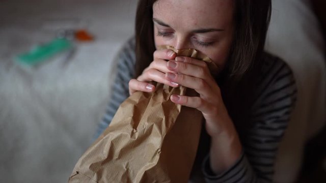Frightened young girl having suddenly panic attack staying at home, fear and lack of air, deep breathing into brown paper bag to calm down. Brain disorder, anxiety and panic shock