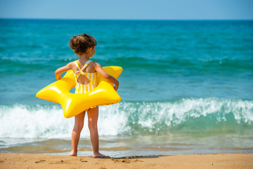 A little child girl 3 years old stands with a yellow swimming ring in the form of a starfish and looks at the sea. Back view