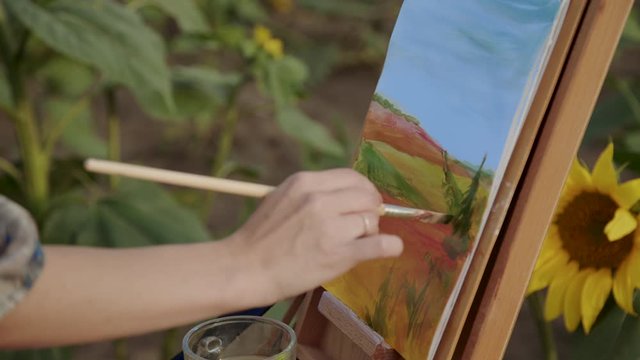Female hand painting a landscape with brush and watercolors among sunflowers
