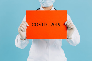 Doctor woman with surgical mask pointing to red paper with message Coronavirus, isolated on blue background