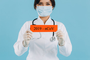 Doctor woman with surgical mask pointing to red paper with message Coronavirus, isolated on blue background