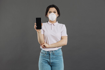 Woman in protective respiratory holding phone and showing something. Coronavirus concept and counteraction.