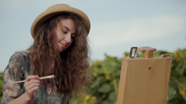 Cheerful long-haired lady painting landscape with watercolors among sunflowers