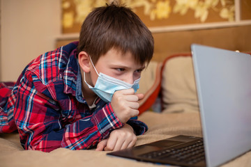 teenage boy in protective medical mask coughs in fist. child remotely does lessons lying on bed near laptop. Online training during coronavirus quarantine