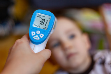 A hand with an electronic thermometer is aimed at the child, on the thermometer the temperature is 38.7, measurement of body temperature by an electronic thermometer