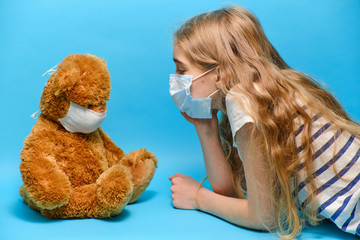 masked blonde girl talking to masked teddy bear. quarantined children concept. 