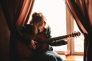 Young woman playing guitar while sitting on windowsill at home