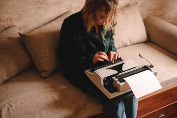 Fototapeta na wymiar Young female writer using typewriter while sitting on sofa at home
