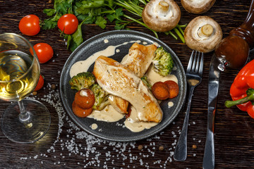Fried chicken fillet in a creamy sauce with broccoli on a dark plate. Tomatoes, mushrooms, fresh herbs, bell peppers and seasonings.