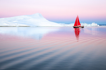 Sail boat with red sails cruising among ice bergs after sunset. Disko Bay, Greenland.