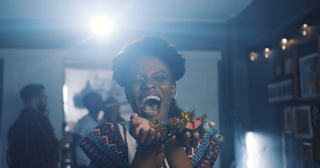 Beautiful happy young African woman laughing, blowing on confetti at fun atmospheric celebration...