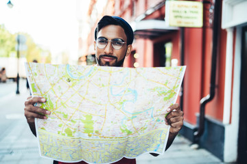 Satisfied ethnic male traveler in stylish outfit reading map at city street
