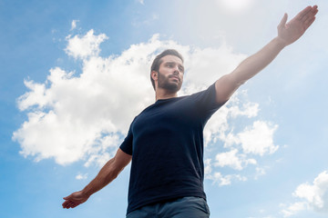 A man wearing a black shirt and dark green shorts stretches his arms out wide in a warrior pose hoping to correct his body in front of a bright blue sky with gorgeous white clouds behind his body.