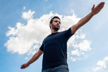Fototapeta na wymiar A man wearing a black shirt and dark green shorts stretches his arms out wide in a warrior pose hoping to correct his body in front of a bright blue sky with gorgeous white clouds behind his body.