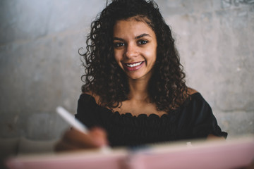 Portrait of cheerful dark skinned curly female student learning and doing homework indoors enjoying education,smiling 20s charming african american hipster girl making notes and looking at camera