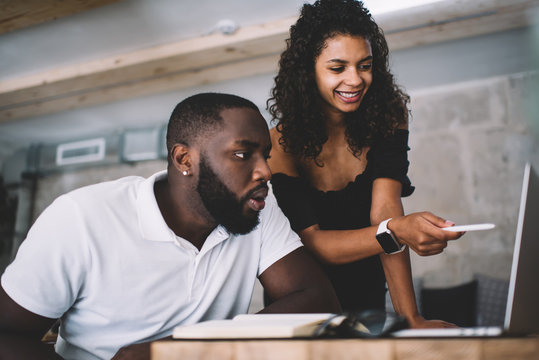 Smiling Dark Skinned Female 20s Explaining Information To Male Student Pointing On Web Page On Laptop Computer, Surprised African American Man Amazed With Browsed Video Communicating With Girlfriend